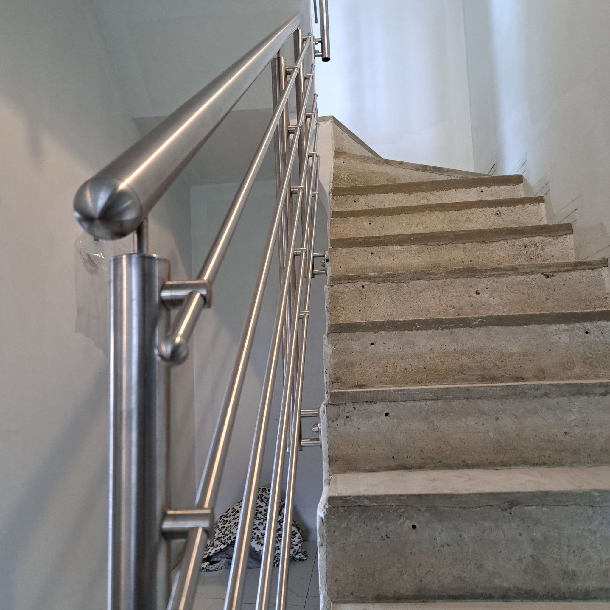Betonowe schody z zamontowaną metalową, nowoczesną barierką ze stali nierdzewnej z kulistym zakończeniem poręczy. W tle biała ściana i fragment podłogi z leżącymi ubraniami.