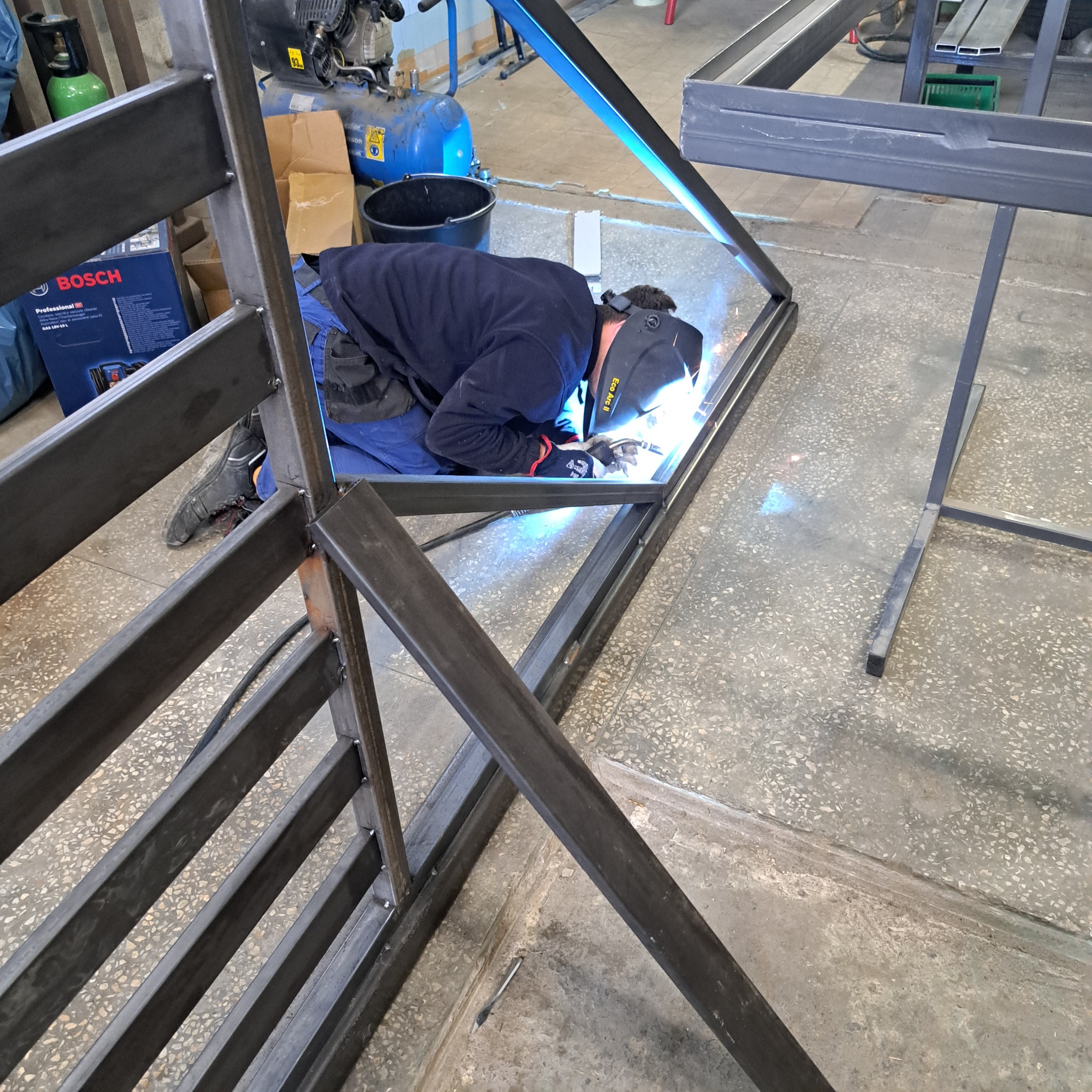 Pracownik w masce spawalniczej łączy metalowe elementy konstrukcji przyszłej balustrady w warsztacie, widoczny iskrzący się łuk elektryczny.