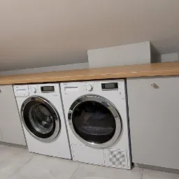 Pralka i suszarka Beko w zabudowie z szarymi szafkami z drewnianym blatem w pomieszczeniu ze skośnym sufitem.