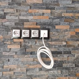 Marcin Żyżniewski - Cztery gniazdka elektryczne, trzy białe i jedno kwadratowe białe z podłączonym białym kablem, zamontowane na ścianie wyłożonej dekoracyjną okładziną kamienną w odcieniach szarości i brązu.