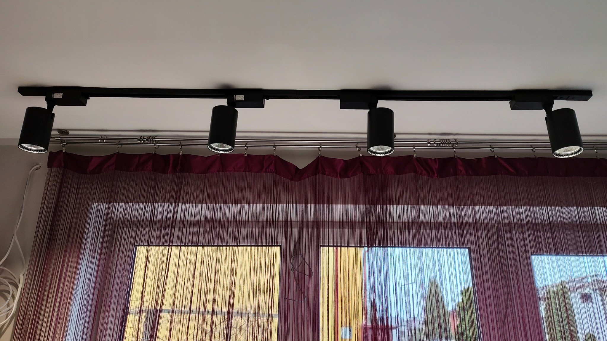 Cztery czarne reflektory szynowe zamontowane na suficie, widok z dołu, z zasłonami sznurkowymi w tle i widokiem z okna.