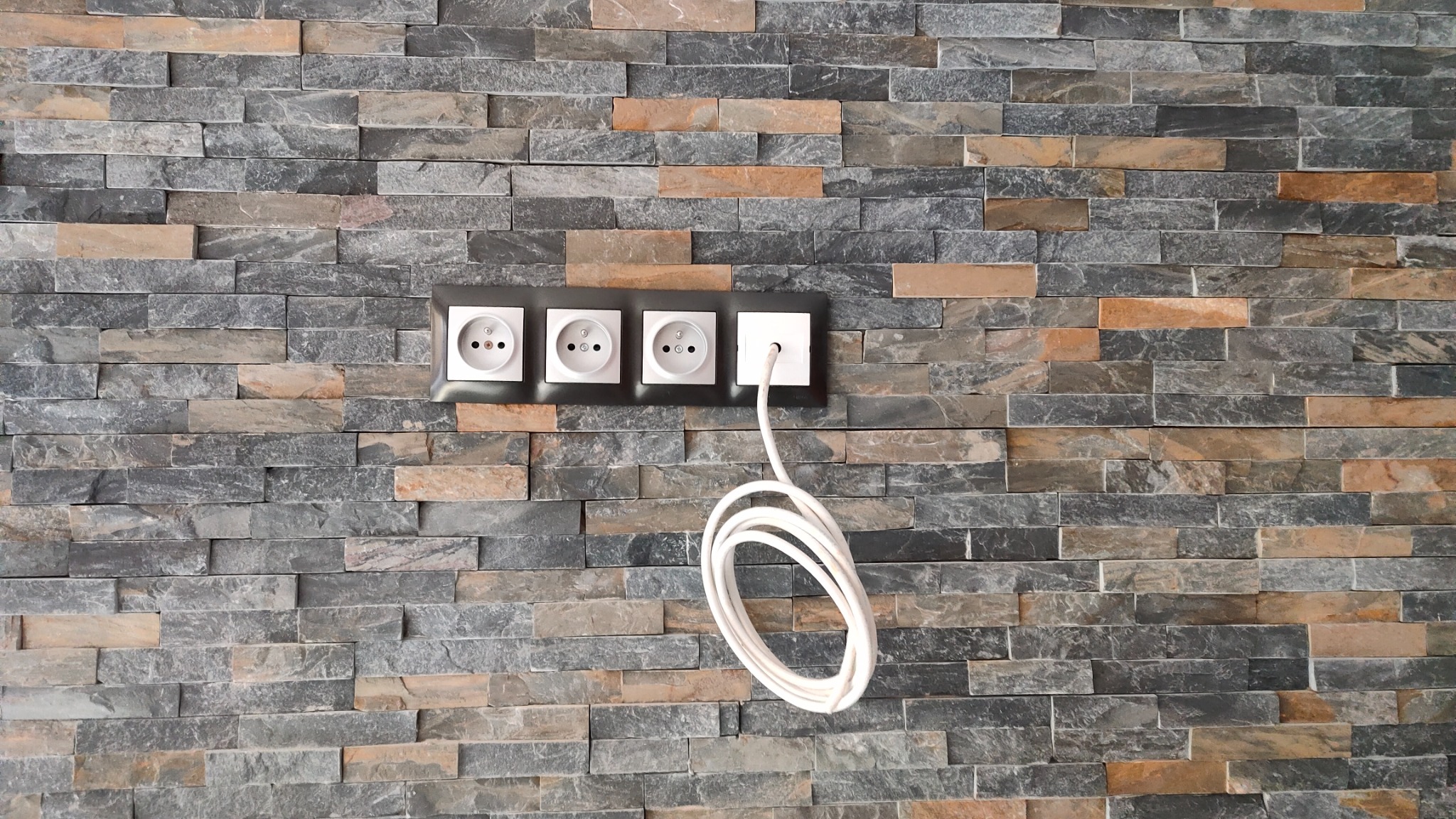 Cztery gniazdka elektryczne, trzy białe i jedno kwadratowe białe z podłączonym białym kablem, zamontowane na ścianie wyłożonej dekoracyjną okładziną kamienną w odcieniach szarości i brązu.