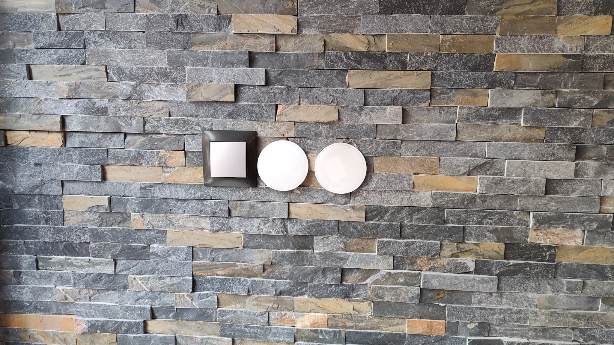 Ściana wyłożona szarymi i beżowymi kamieniami z zamontowanym kwadratowym włącznikiem światła marki Simon oraz dwoma okrągłymi lampami LED.