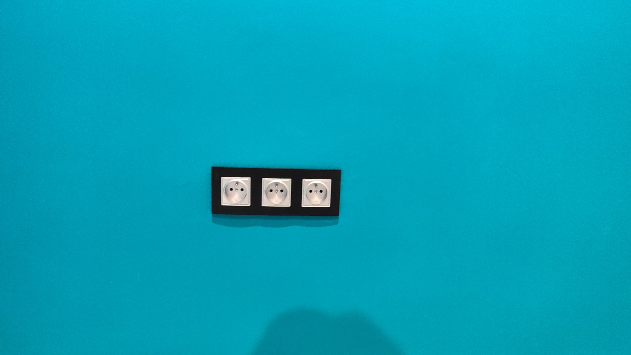 Trzy gniazdka elektryczne z bolcem uziemiającym, zamontowane w czarnej ramce na turkusowej ścianie, widoczne z lekkiego dołu.