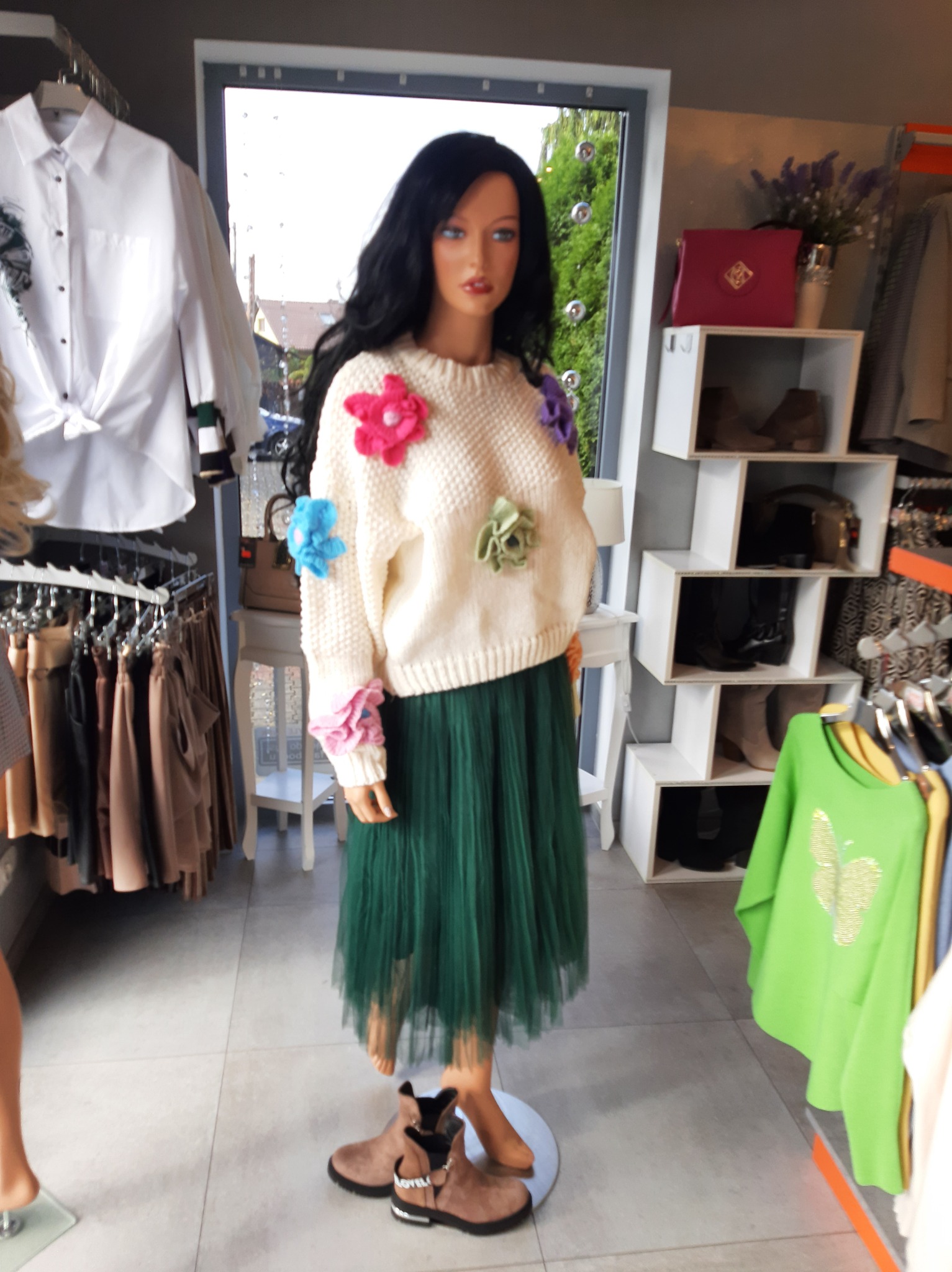 Manekin w kremowym swetrze z aplikacjami kwiatowymi i zielonej tiulowej spódnicy, prezentowany w butiku z różnorodną odzieżą i akcesoriami na wieszakach i półkach.