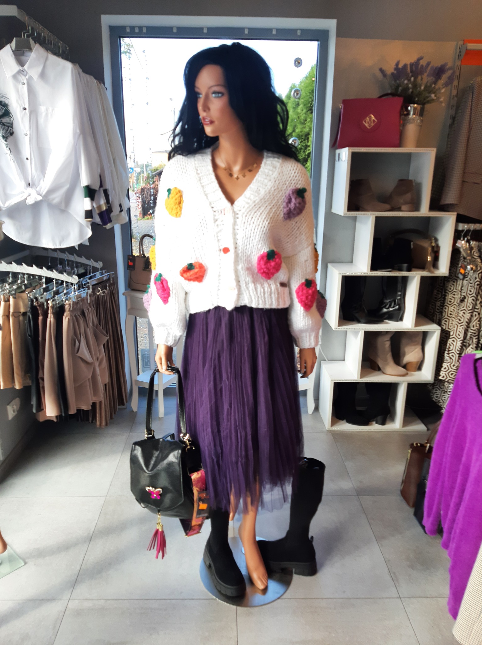 Manekin w Katowicach prezentuje biały sweter z aplikacjami w kształcie owoców, fioletową tiulową spódnicę i czarne botki, trzymając czarną torebkę z różowym akcentem.