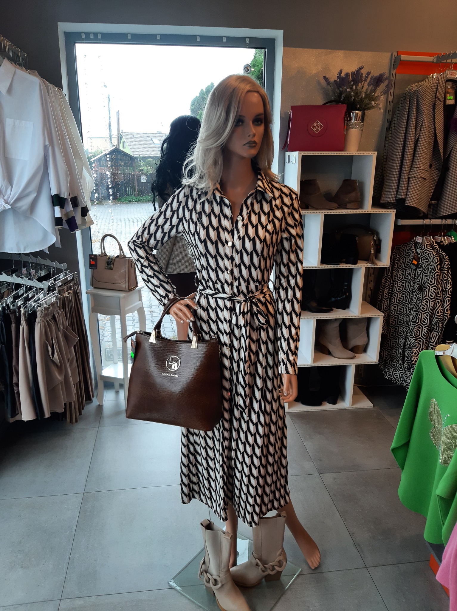 Manekin w czarno-białej sukience z geometrycznym wzorem, trzymający brązową torebkę z logo, obok beżowe botki, na tle wnętrze sklepu odzieżowego z różnymi ubraniami.