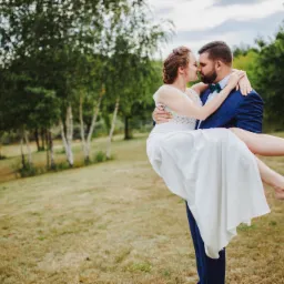 Pan młody w granatowym garniturze trzyma na rękach pannę młodą w białej sukni na tle łąki i drzew.