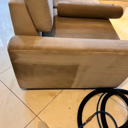 Twórcy Czystości - Fragment beżowej, modułowej sofy z widoczną różnicą w czystości po fragmencie wyczyszczonej tapicerki, obok leży dysza odkurzacza piorącego z wężami na podłodze w jasne płytki.