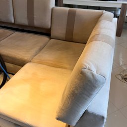 Twórcy Czystości - Beżowa, narożna kanapa z widocznymi plamami na siedzisku, sugerująca potrzebę odświeżenia, z fragmentem węża odkurzacza widocznym w lewym dolnym rogu i elementami wyposażenia mieszkania w tle.