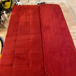 Twórcy Czystości - Czerwona, rozkładana sofa podczas czyszczenia; widoczna część wyczyszczona i część jeszcze mokra, obok żółty odkurzacz piorący i butelki z detergentami.