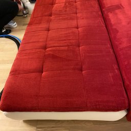 Twórcy Czystości - Czerwona kanapa modułowa z kwadratowymi przeszyciami, częściowo wyczyszczona, z widocznymi śladami wilgoci po praniu, obok odkurzacz piorący Kärcher i butelki ze środkami czyszczącymi.