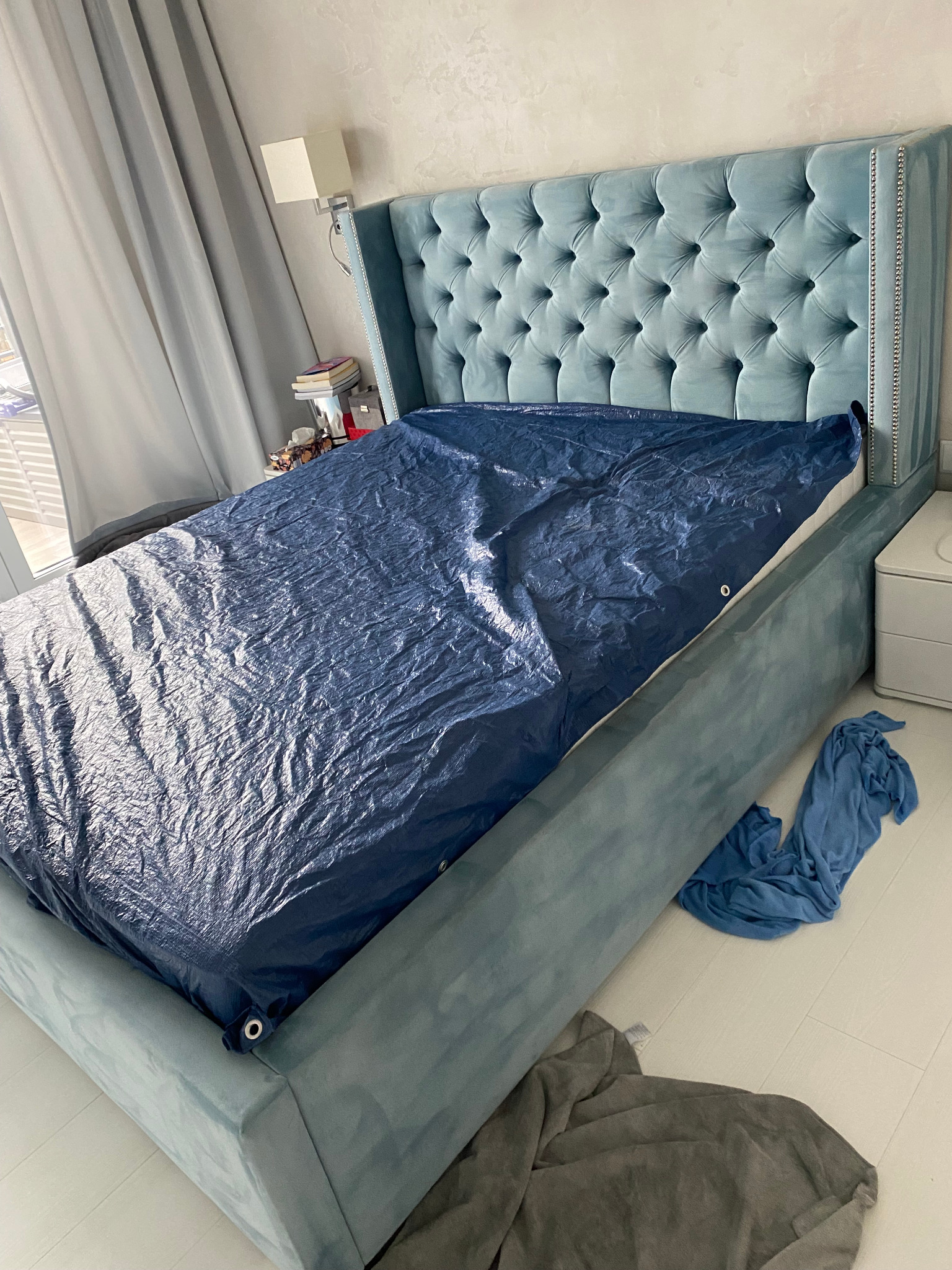 Łóżko z welurowym zagłówkiem zabezpieczone niebieską folią przed zabrudzeniami, prawdopodobnie przed czyszczeniem tapicerki.