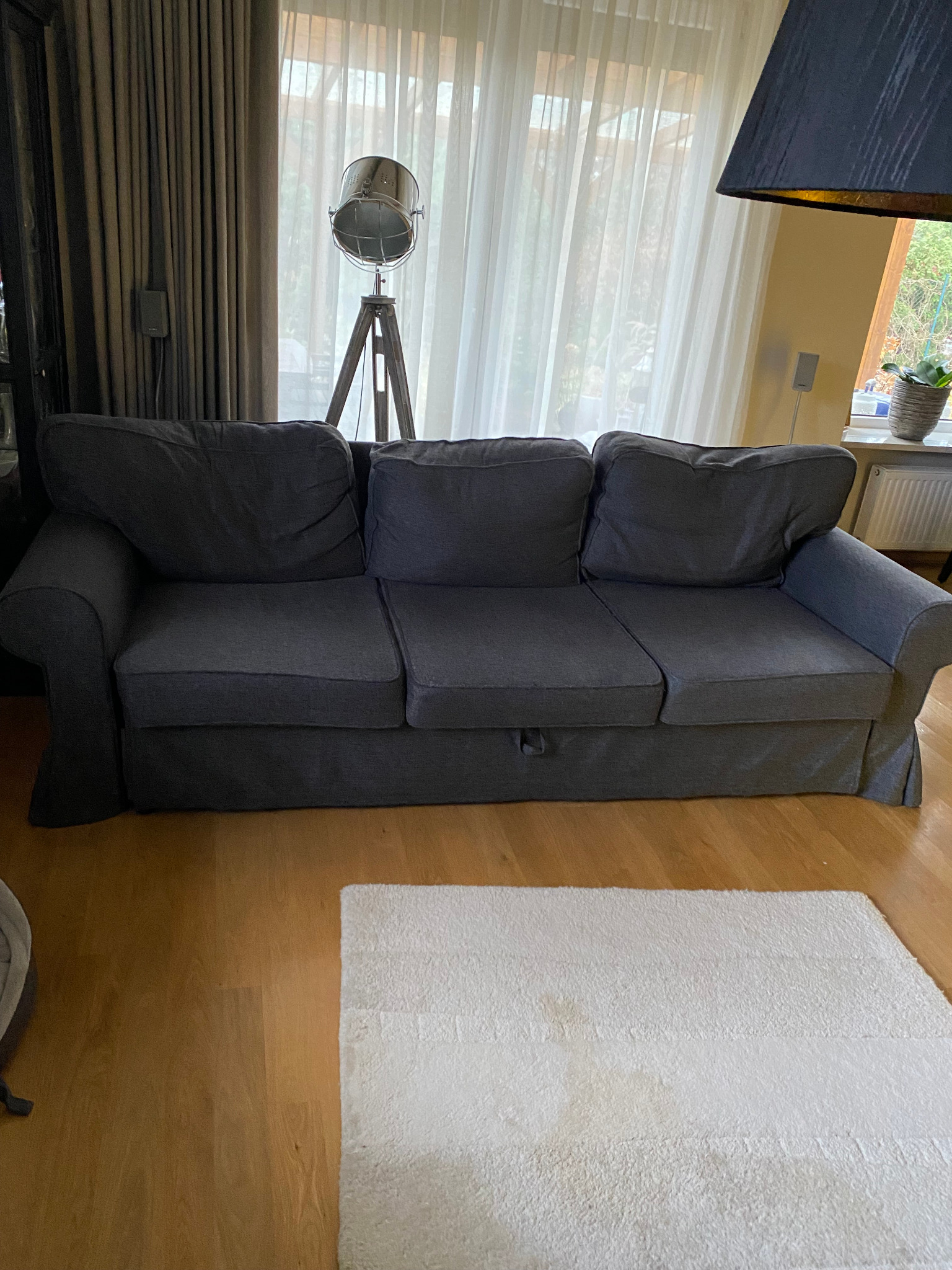 Szara, trzyosobowa sofa z pokrowcem w salonie, widoczna na tle okna z zasłonami i lampy stojącej. Pod sofą leży biały dywan.