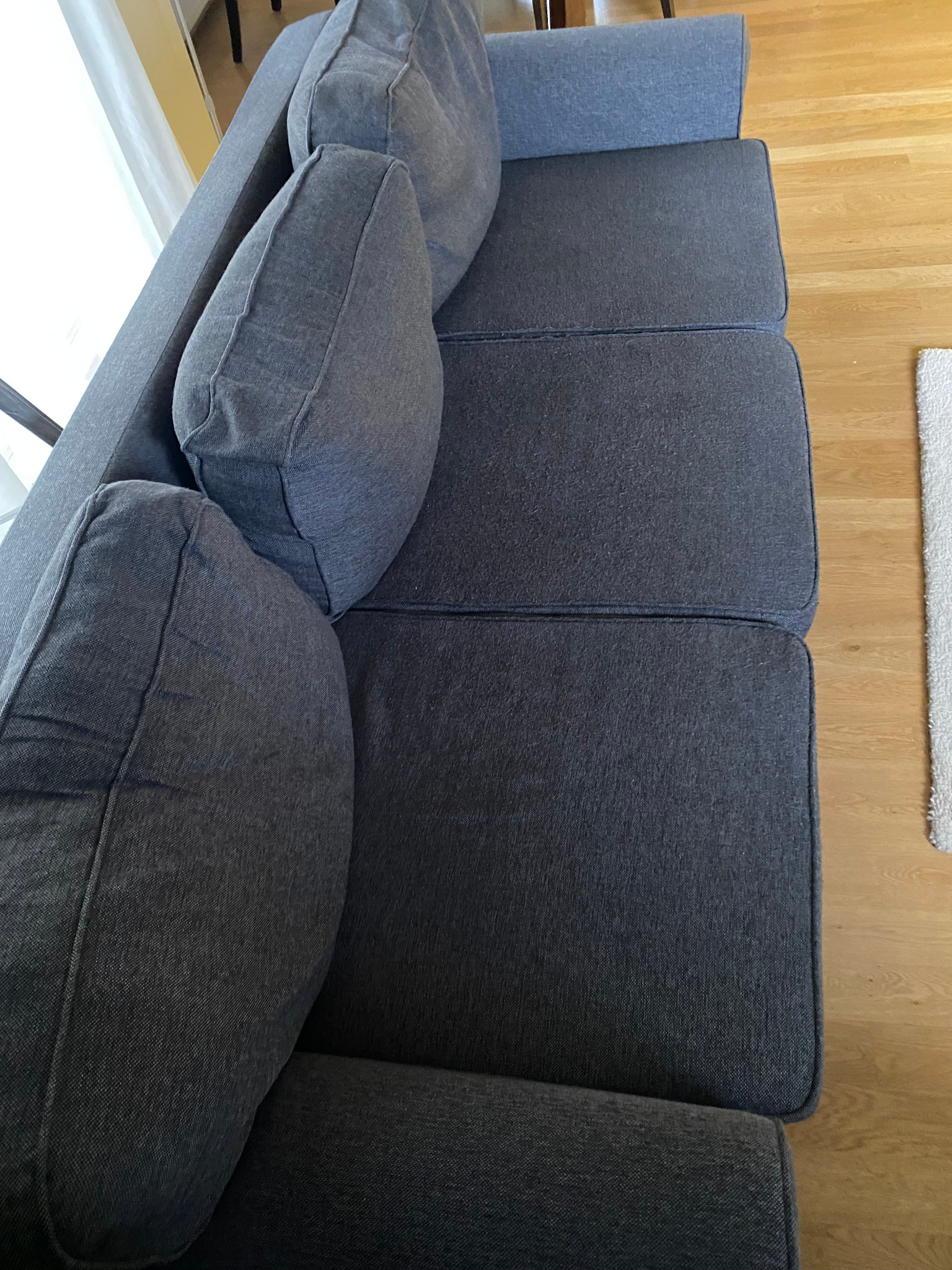 Szara, tapicerowana sofa z poduszkami, widok z góry, na drewnianej podłodze, obok biały dywanik.