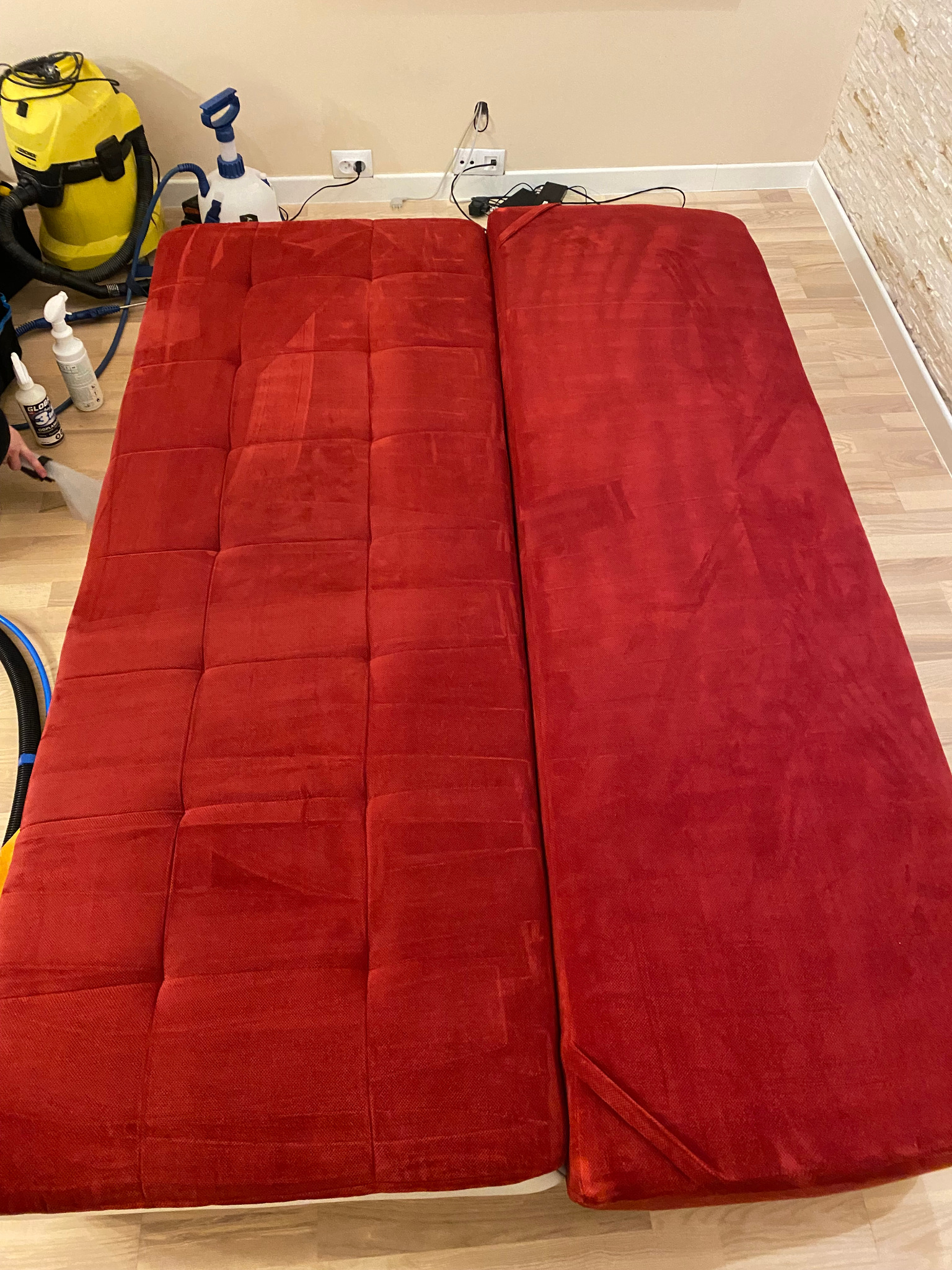 Czerwona, rozkładana sofa podczas czyszczenia; widoczna część wyczyszczona i część jeszcze mokra, obok żółty odkurzacz piorący i butelki z detergentami.