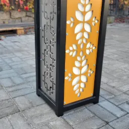 Nowoczesna, metalowa osłona na donicę w kolorze czarnym, z ażurowymi panelami w kolorze szarym i pomarańczowym z motywem roślinnym, stojąca na brukowanej nawierzchni, z jesiennym tłem.