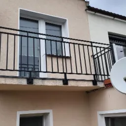 Czarna, metalowa balustrada balkonowa z pionowymi szczeblami, zamontowana na balkonie budynku o beżowej elewacji, widoczna antena satelitarna na sąsiedniej ścianie.