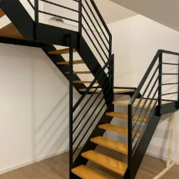 Nowoczesne schody wewnętrzne z czarną, metalową konstrukcją i drewnianymi stopniami, widoczne z dołu, z barierką z poziomych prętów i fragmentem antresoli w tle.