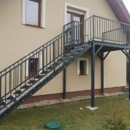 Zewnętrzne schody stalowe z balustradą prowadzące na balkon, zamontowane przy beżowym budynku z brązowymi oknami i drzwiami balkonowymi.