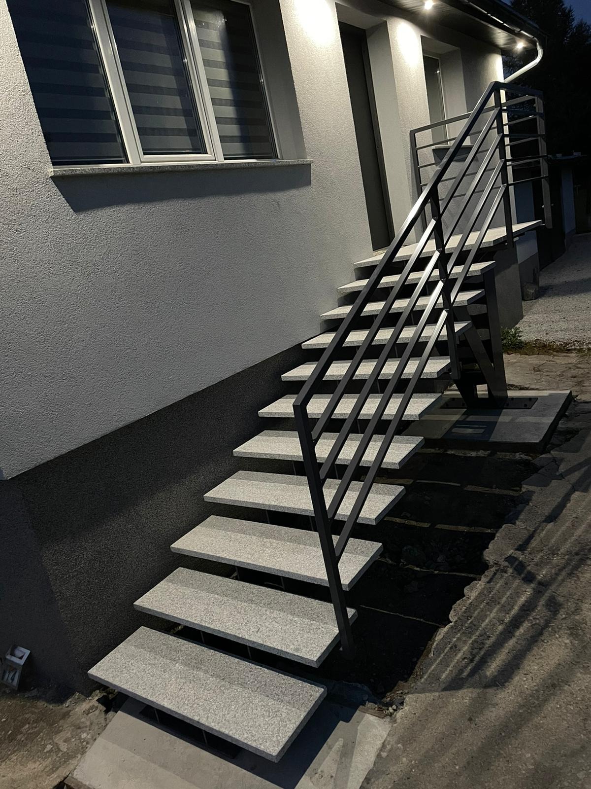 Zewnętrzne schody z szarymi, granitowymi stopniami i czarną, metalową balustradą o minimalistycznym designie, oświetlone punktowymi lampami, przylegające do szarego budynku.