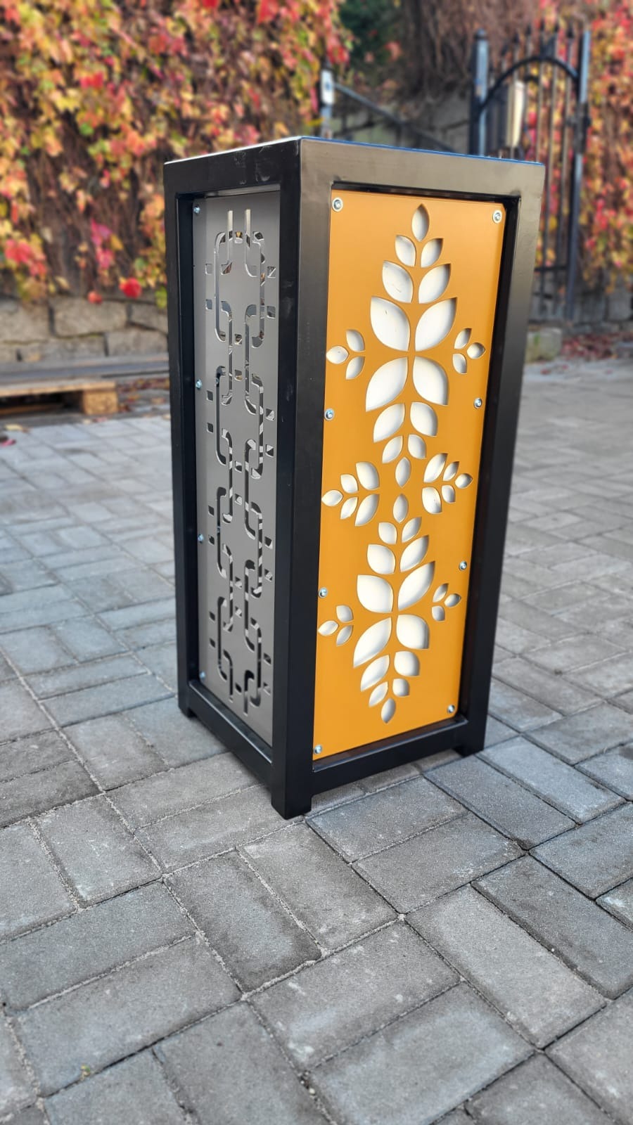 Nowoczesna, metalowa osłona na donicę w kolorze czarnym, z ażurowymi panelami w kolorze szarym i pomarańczowym z motywem roślinnym, stojąca na brukowanej nawierzchni, z jesiennym tłem.