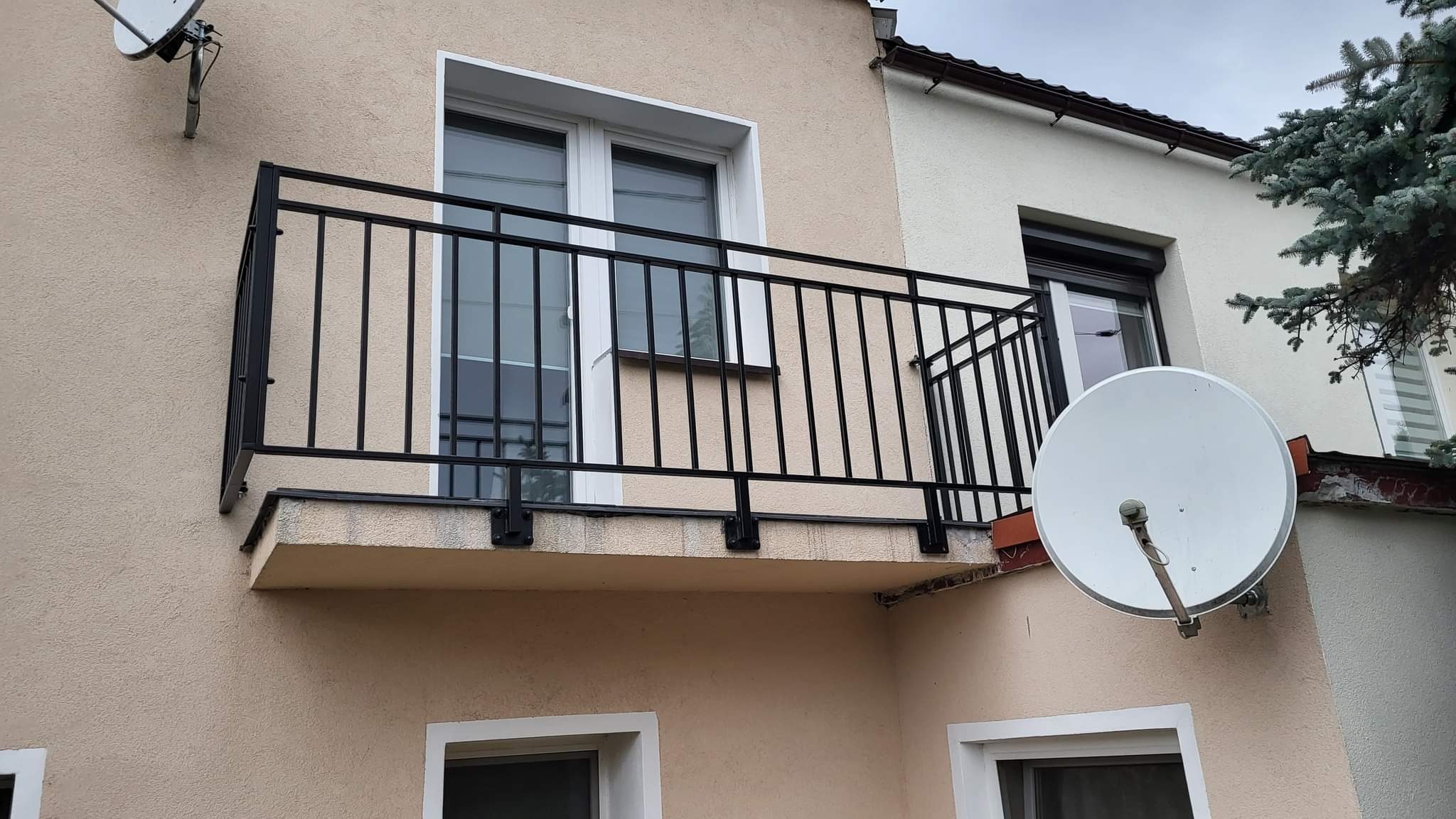 Czarna, metalowa balustrada balkonowa z pionowymi szczeblami, zamontowana na balkonie budynku o beżowej elewacji, widoczna antena satelitarna na sąsiedniej ścianie.
