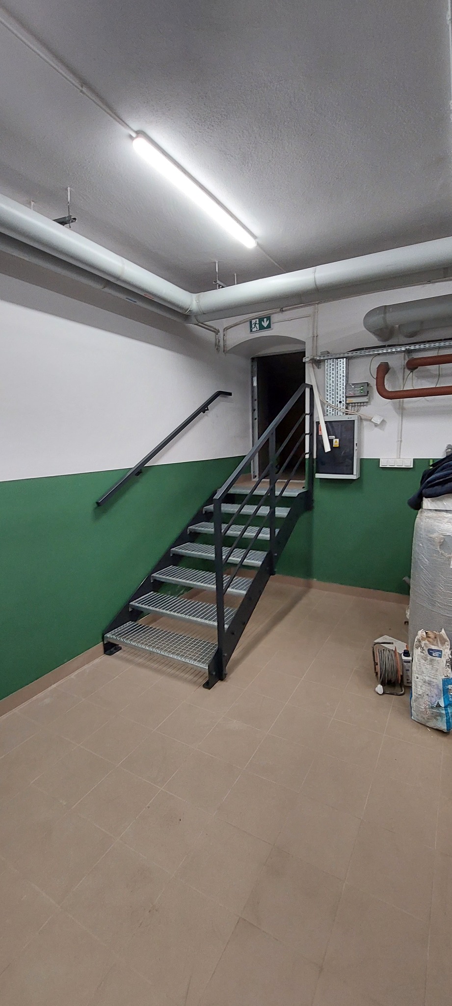 Metalowe schody z ażurowymi stopniami i czarną poręczą w piwnicy z zielono-białymi ścianami, prowadzące do ciemnego przejścia.