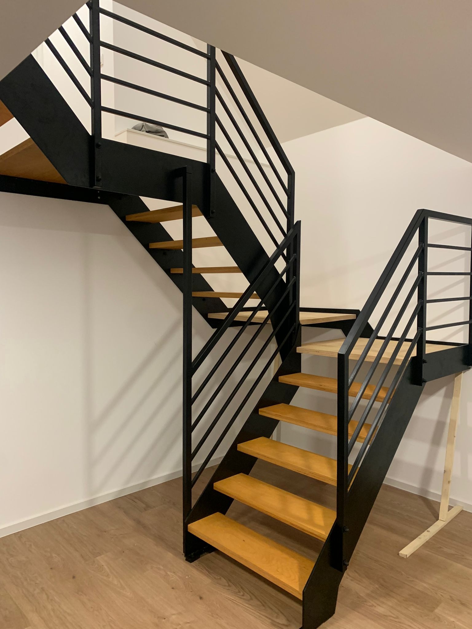 Nowoczesne schody wewnętrzne z czarną, metalową konstrukcją i drewnianymi stopniami, widoczne z dołu, z barierką z poziomych prętów i fragmentem antresoli w tle.