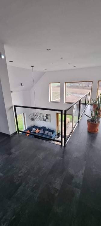 Wnętrze domu z czarną podłogą i szklaną balustradą z czarnymi ramami, widok z góry na salon z szarą sofą i kolorowymi poduszkami.