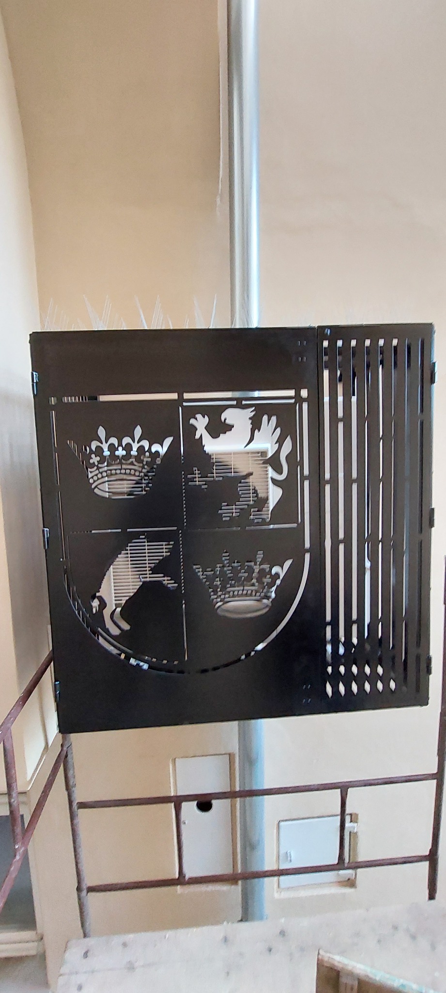 Metalowa osłona z wyciętym herbem (korony, lew, ptak) zamontowana na balkonie, z kolcami przeciw ptakom na górnej krawędzi.