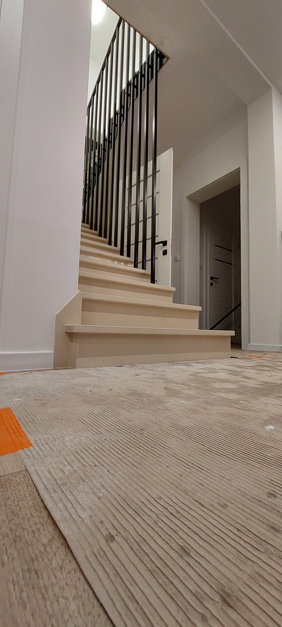 Wnętrze domu w trakcie remontu z widocznymi schodami z beżowego kamienia i czarną, minimalistyczną balustradą. Na podłodze widoczne płyty kartonowo-gipsowe zabezpieczające podłogę.