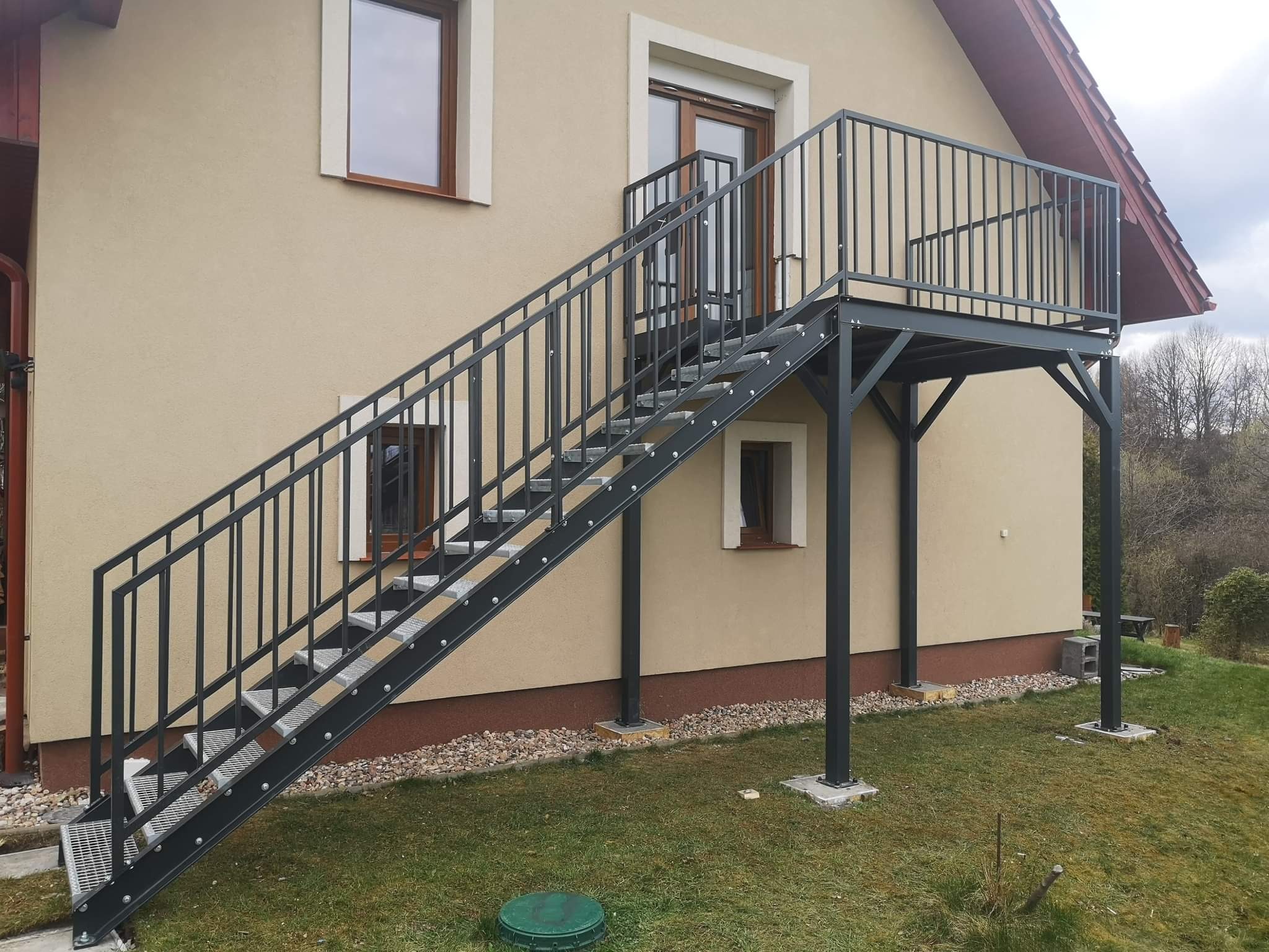 Zewnętrzne schody stalowe z balustradą prowadzące na balkon, zamontowane przy beżowym budynku z brązowymi oknami i drzwiami balkonowymi.