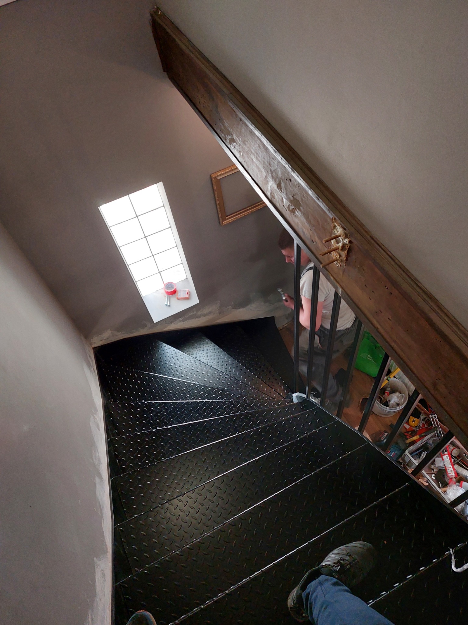 Montaż czarnych, metalowych schodów z blachy ryflowanej wewnątrz budynku, widok z góry na pracownika podczas mocowania balustrady z metalowych prętów, na podeście widoczne narzędzia.