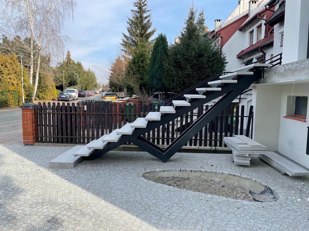 Zewnętrzne schody z czarną metalową konstrukcją i jasnoszarymi stopniami, w trakcie montażu, z widocznymi płytami do wykończenia terenu.