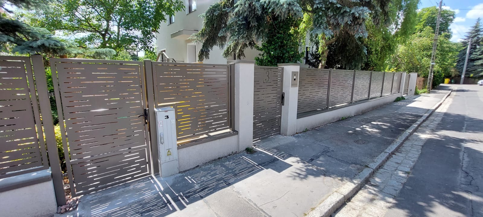 Nowoczesne ogrodzenie z metalowymi panelami z poziomymi wycięciami, brama z numerem domu '3', szara kolorystyka, betonowe słupki, widok z ulicy w słoneczny dzień.