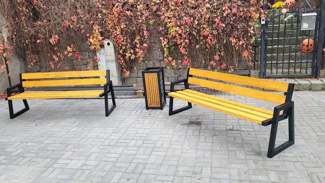 Dwie ławki parkowe z żółtego drewna i czarnymi metalowymi nogami, stojące na brukowanej nawierzchni, z koszem na śmieci w tle i dekoracjami halloweenowymi na bramie.