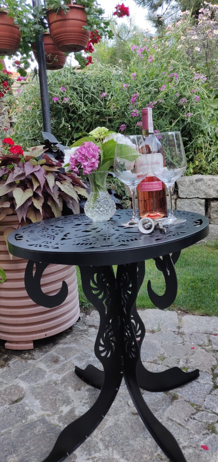 Okrągły, czarny stolik metalowy z ażurowym blatem i dekoracyjnymi nogami w ogrodzie, na którym stoi butelka różowego wina, dwa kieliszki, korkociąg i wazon z kwiatami.