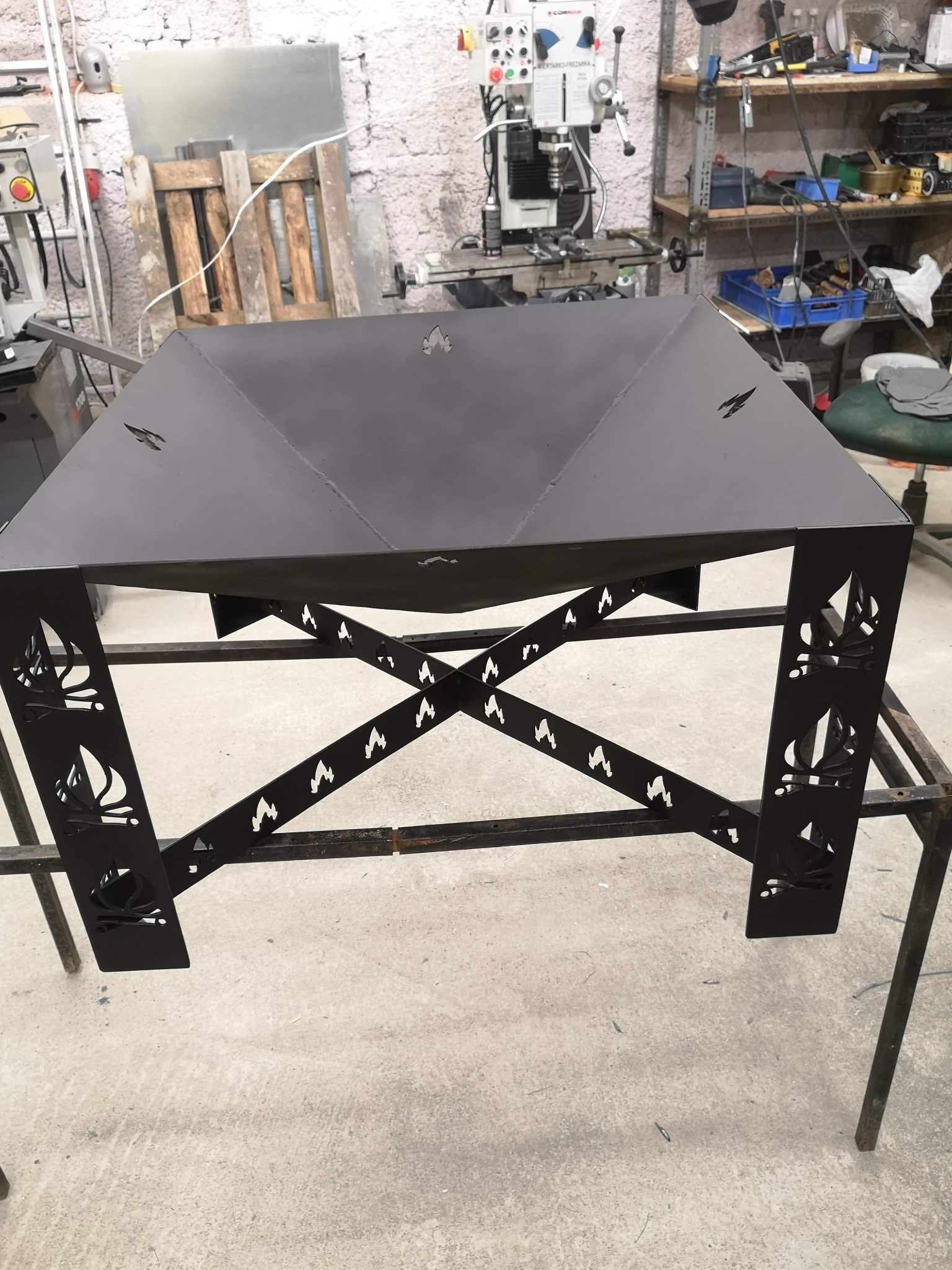 Czarny, metalowy kosz na ognisko z wyciętymi motywami ognia i żaglówek, stojący na warsztatowym stole, w tle widoczna ściana z cegły i maszyny warsztatowe.