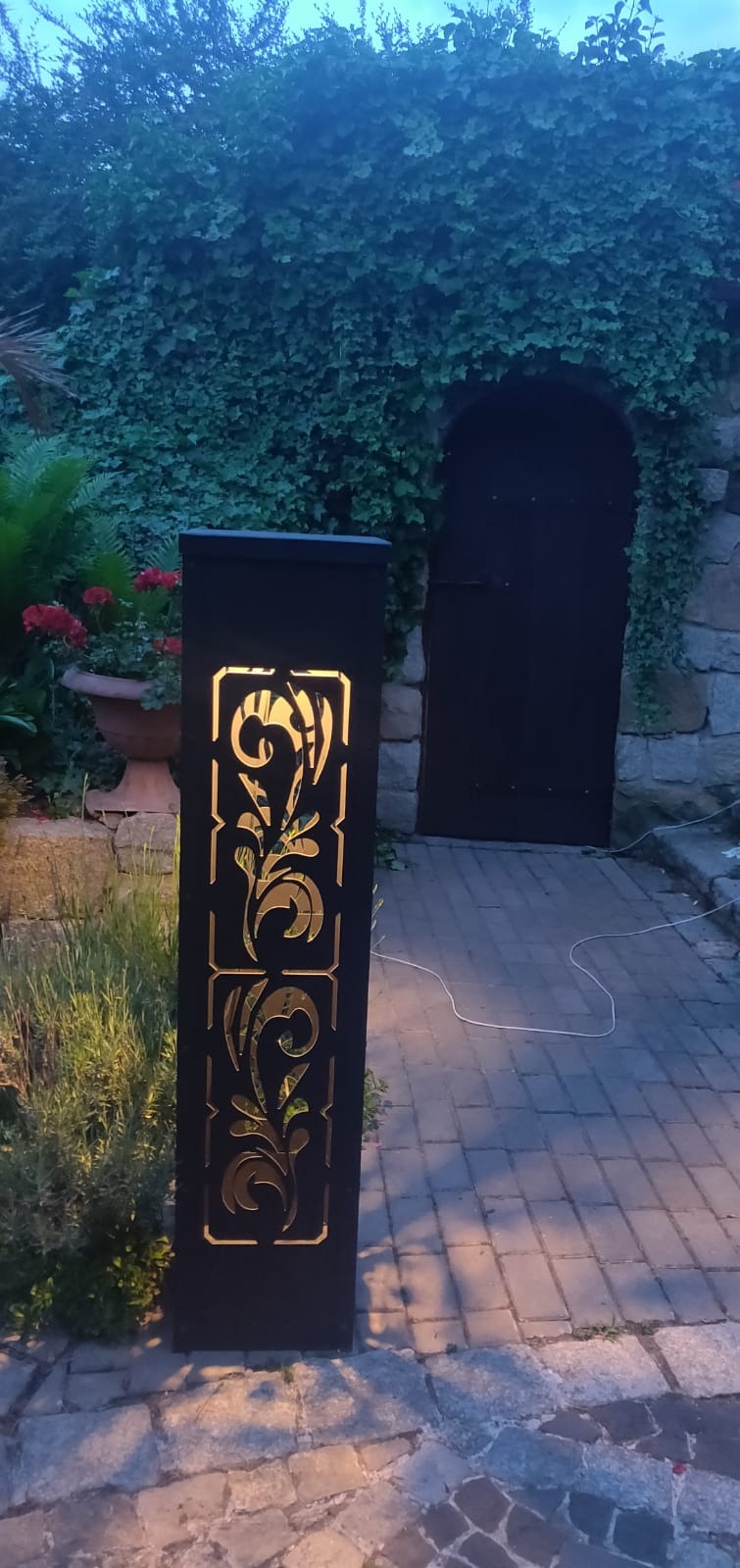 Ozdobna, podświetlana lampa ogrodowa z wycinanym wzorem roślinnym, stojąca na brukowanej ścieżce, z kamiennym murem i porośniętymi bluszczem drzwiami w tle.