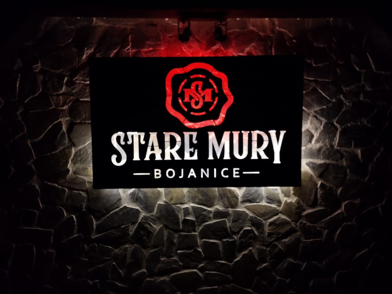 Podświetlany szyld reklamowy z logo i nazwą 'Stare Mury - Bojanice' na tle kamiennej ściany, oświetlony od góry czerwonym światłem.