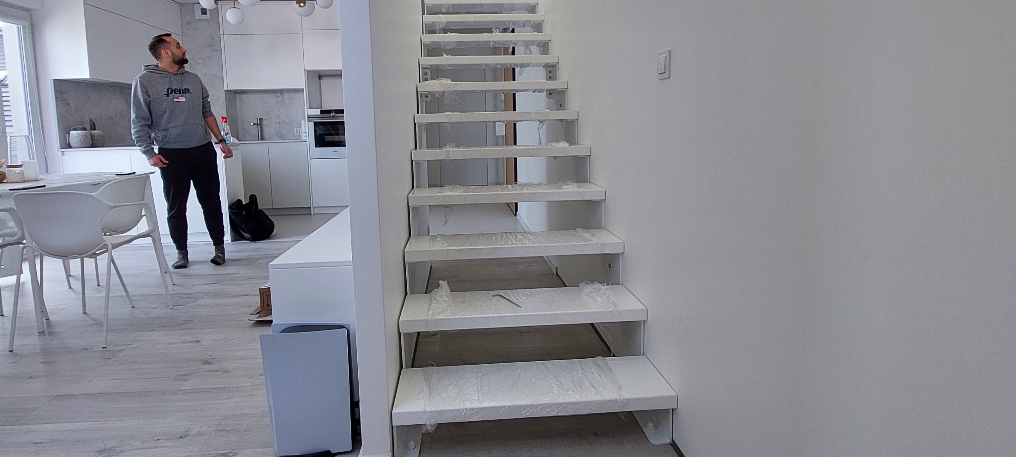 Nowoczesne schody ażurowe, białe, z zabezpieczeniami z folii, w jasnym wnętrzu domu z widoczną kuchnią i mężczyzną w tle.