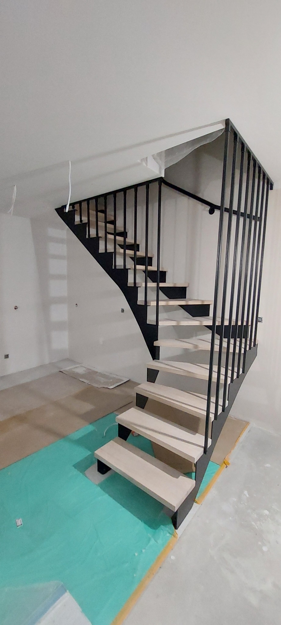 Schody wewnętrzne z drewnianymi stopniami i czarną, metalową balustradą w surowym wnętrzu, zabezpieczonym folią malarską.