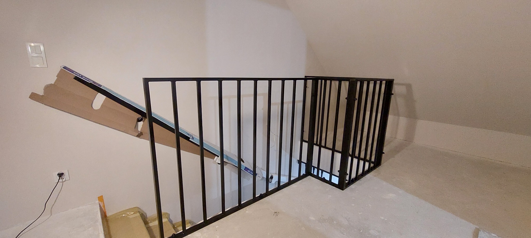 Czarna, metalowa balustrada schodowa z pionowymi prętami, zamontowana przy schodach w trakcie remontu, z widocznymi materiałami budowlanymi i kartonami na podłodze.