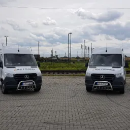 Dwa białe busy marki Mercedes z logo Rktrans i chromowanymi orurowaniami z przodu, zaparkowane na placu z kostki brukowej, w tle tory kolejowe i cysterny.