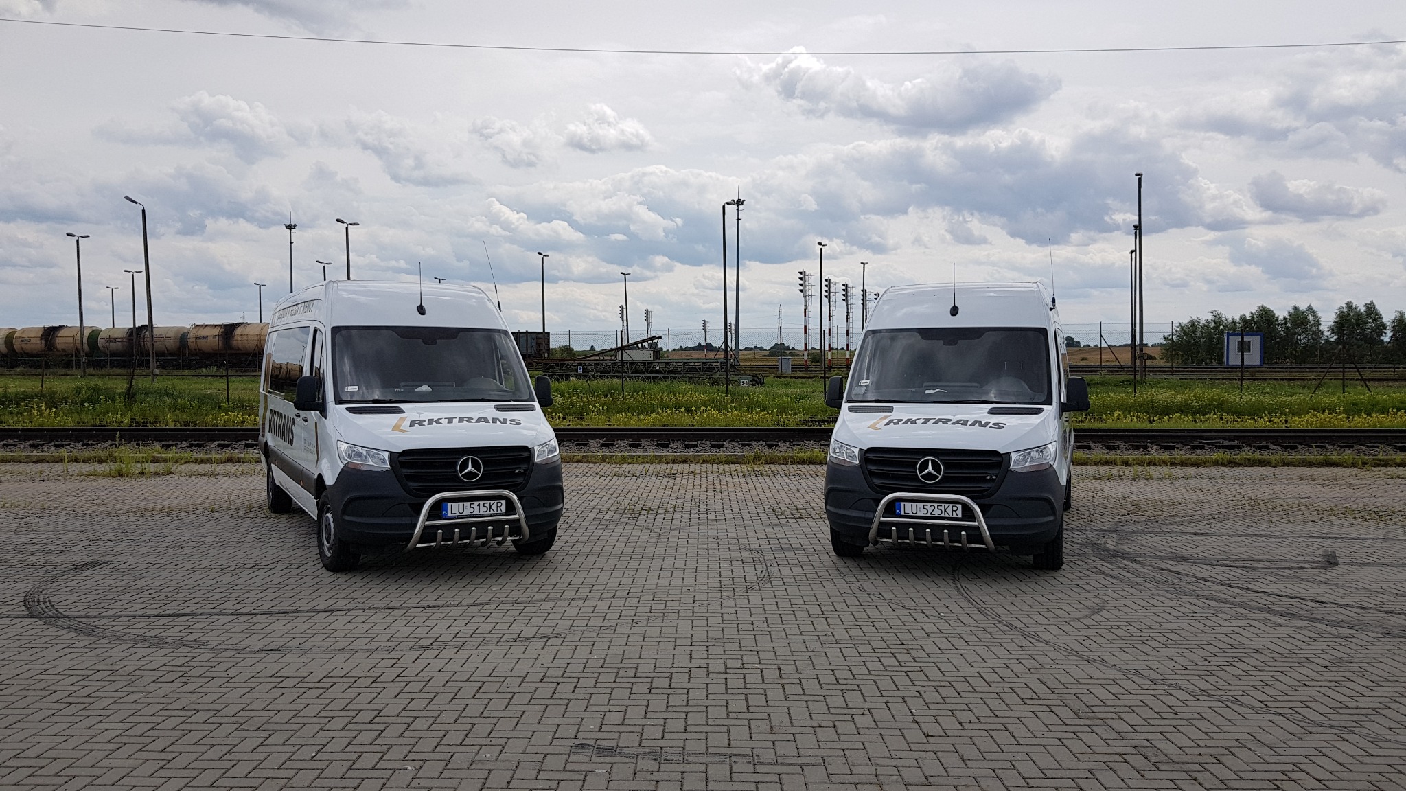 Dwa białe busy marki Mercedes z logo Rktrans i chromowanymi orurowaniami z przodu, zaparkowane na placu z kostki brukowej, w tle tory kolejowe i cysterny.