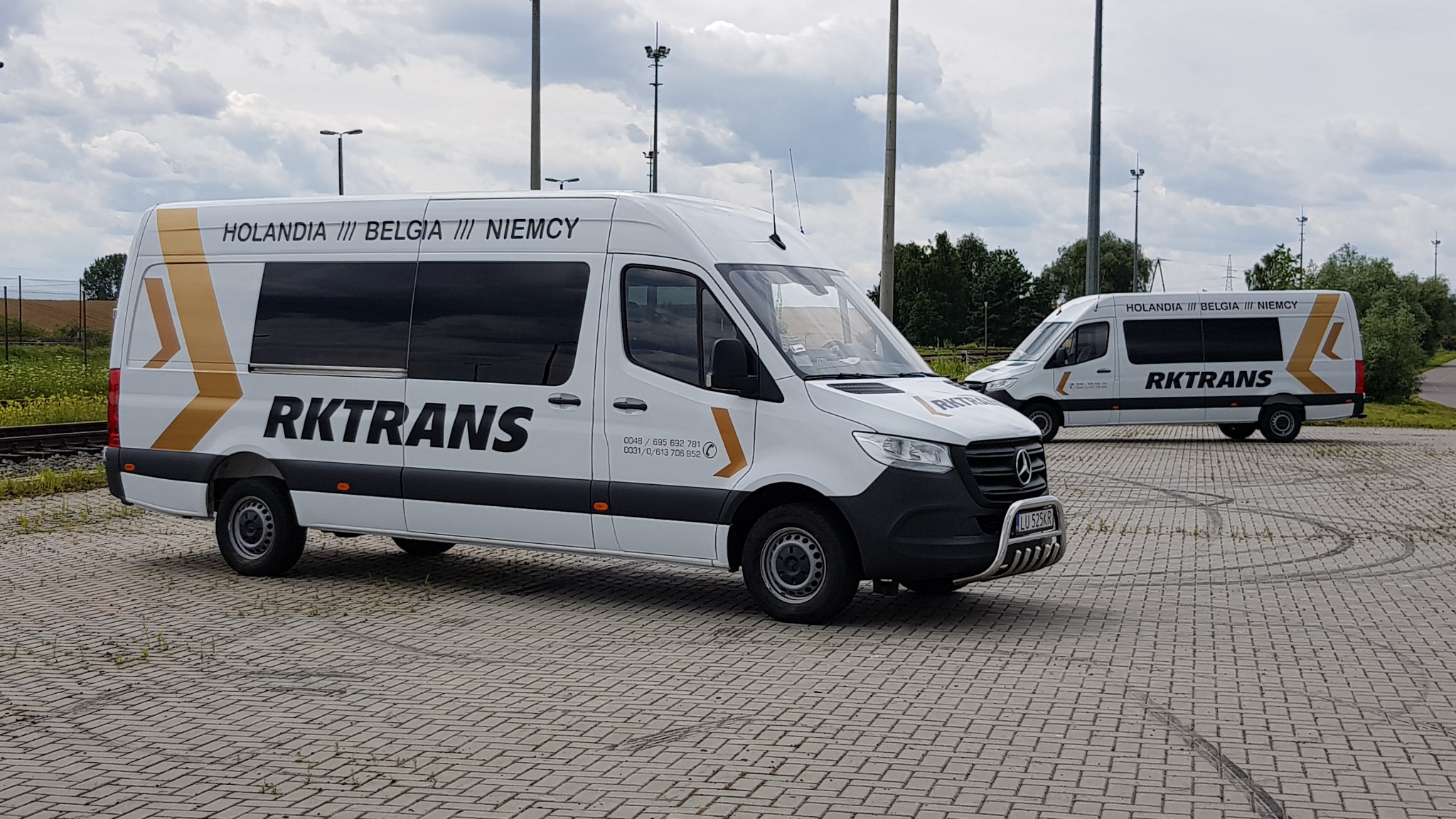 Dwa białe busy z logo Rktrans, napisem 'Holandia/Belgia/Niemcy' i złotymi strzałkami na karoserii, zaparkowane na placu wyłożonym kostką brukową w pochmurny dzień.