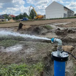 Testowanie wydajności studni: strumień wody wypływa z niebieskiej rury zakończonej metalową armaturą na tle placu budowy z betoniarką i nowym budynkiem.