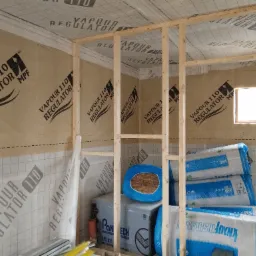 Wnętrze domu szkieletowego w trakcie budowy: widoczne drewniane ramy ścian, izolacja w rolkach Knauf Insulati i folia paroizolacyjna Vapour 110 Regulator na ścianach.