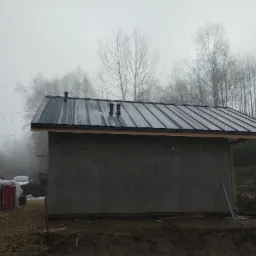 Niewielki budynek w trakcie budowy z czarnym metalowym dachem i surowymi ścianami, w tle mglisty las i materiały budowlane.